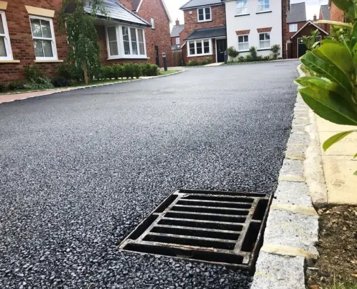 A newly paved driveway with a drain grate is surrounded by brick houses whilst greenery lines the street.