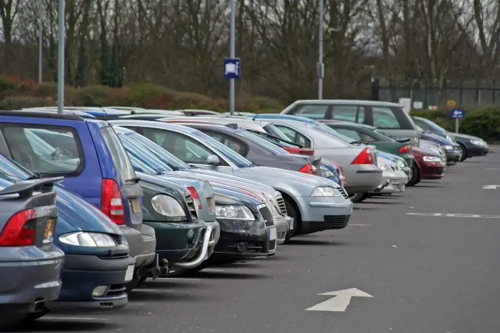 A row of cars parked in an outdoor car park on a cloudy day. Vehicles vary in color and model. Arrow marks guide traffic.
