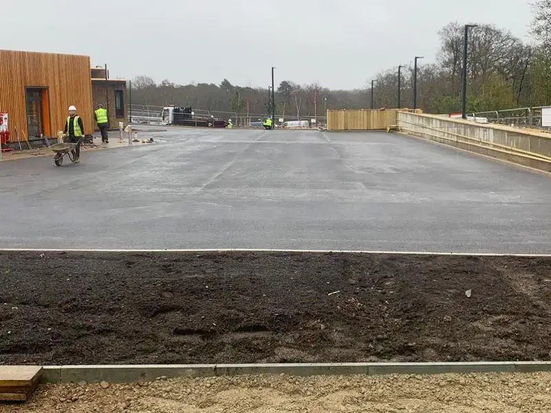 A newly paved car park surrounded by construction workers in yellow vests, with wooden fencing and trees in the background.