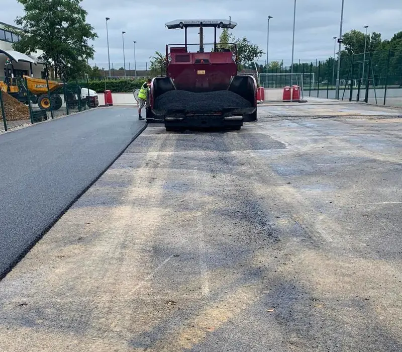 A large paver machine spreads fresh asphalt on a public court, with a worker nearby.