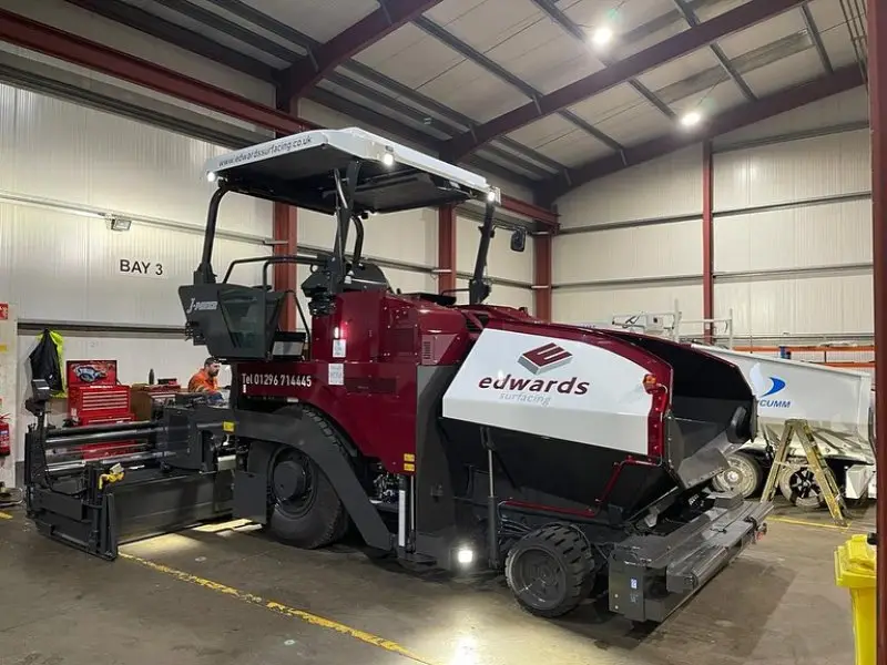 A large, maroon paving machine labeled "Edwards Surfacing" stands in a brightly lit industrial garage, surrounded by tools and equipment.