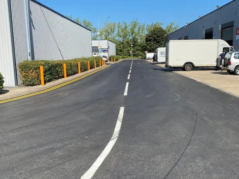 Wide road flanked by industrial buildings, with a white dividing line and parked vehicles on the right.