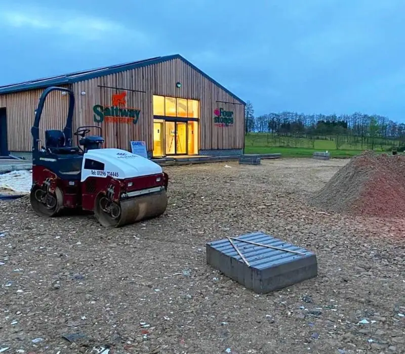 A small road roller sits on gravel near a modern building with lit glass doors, surrounded by construction materials and dirt piles at dusk.