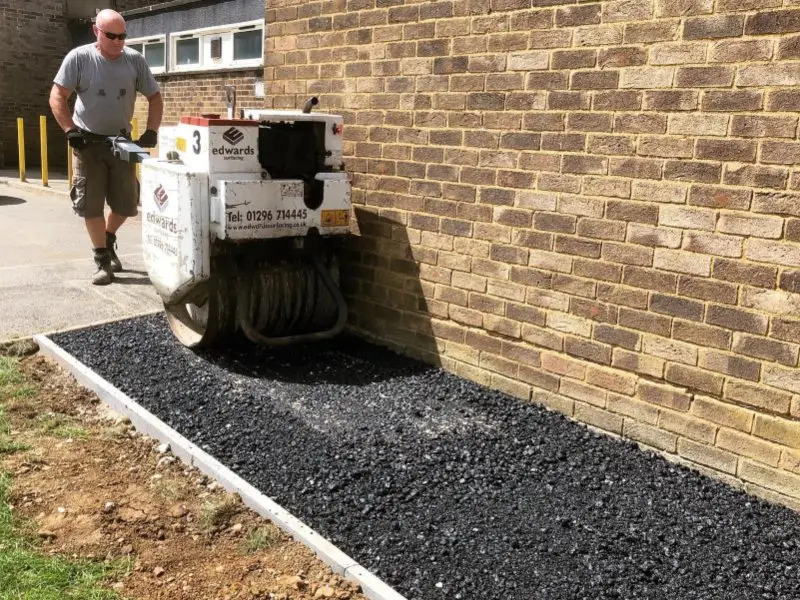 A man operates a small roller compacting asphalt beside a brick wall on a sunny day. The area is freshly paved.