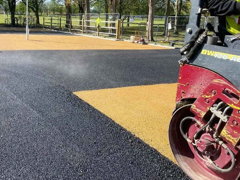 A steamroller compacts a mix of black and yellow tarmac in a sunny park.
