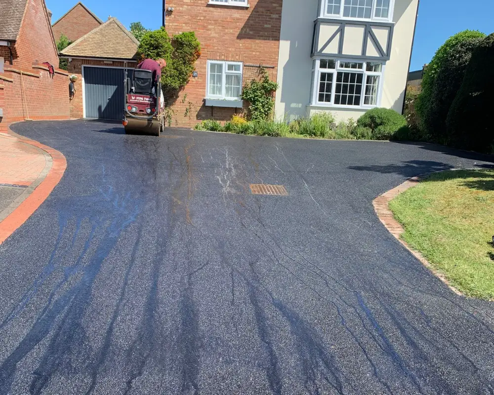 Small road roller compacts freshly laid asphalt on a curved residential driveway in front of a brick house with a bay-windowed facade.