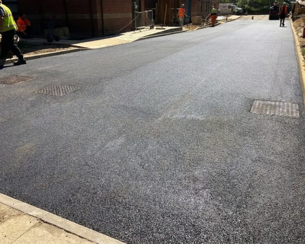 Construction site on a street with fresh asphalt; workers in high-visibility vests along the sidewalk and curb.