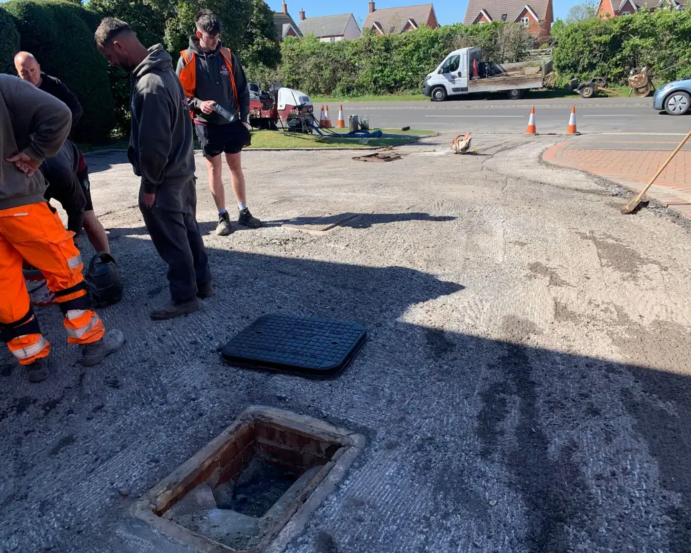 Construction crew works on a damaged road with an open manhole and a removed grate nearby.