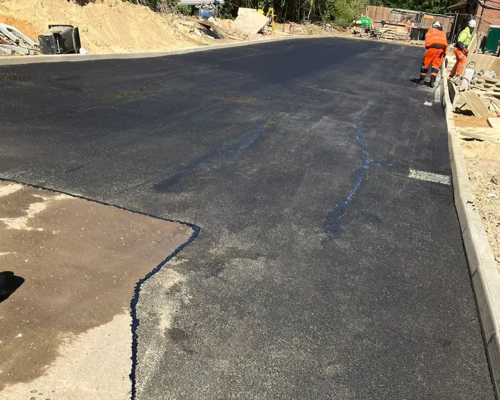 New asphalt road being laid at a construction site; workers in orange safety gear along the curb.