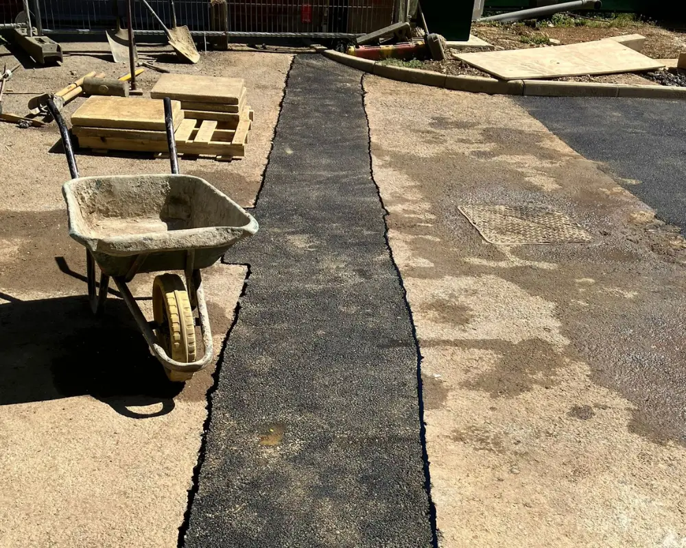 Construction site with a wheelbarrow, wooden pallets, and scattered tools along a paved path under repair.