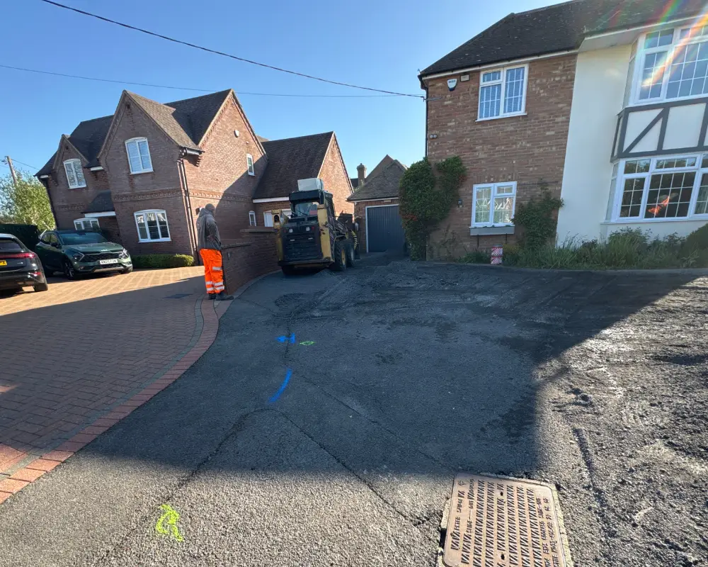 Suburban street with roadwork: a worker in high‑visibility orange trousers stands next to asphalt paving equipment on a freshly laid driveway.