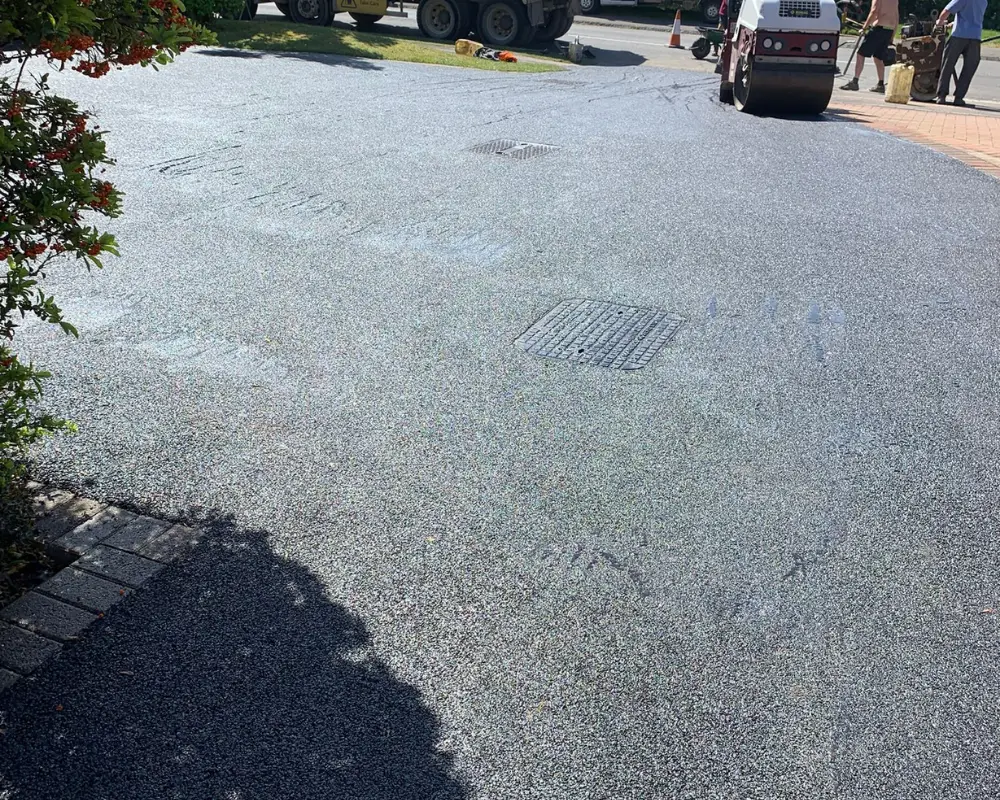 Road construction scene with a roller compactor rolling fresh asphalt, workers and traffic cones in the background.