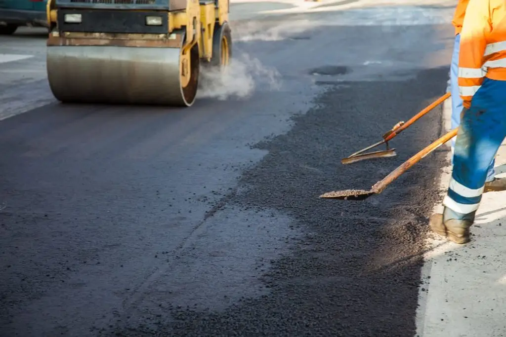 Road workers in high-visibility gear spread and smooth fresh asphalt as a roller compacts the surface ahead of them.