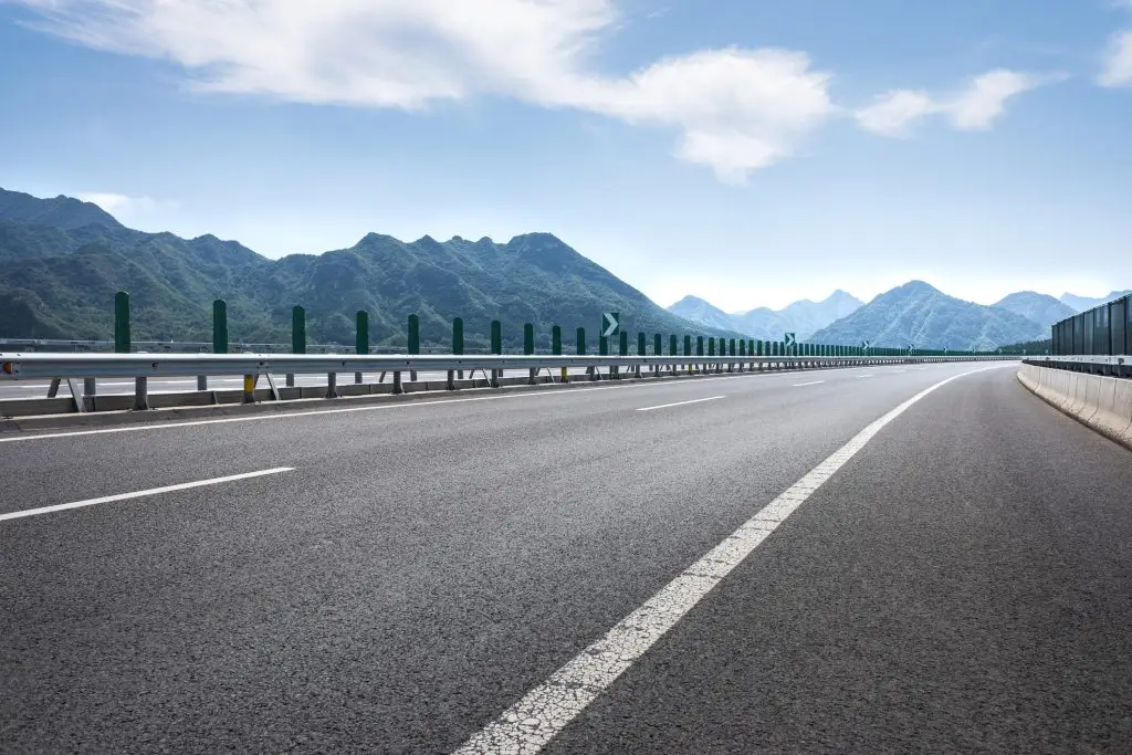 Wide highway scene with a metal guardrail and green posts along the left, mountains in the distance under a clear blue sky; the road curves to the right.